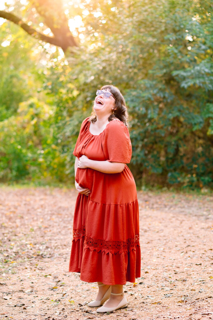 Expectant mother laughing while holding her baby bump during McKinney maternity photos in a wooded outdoor setting.