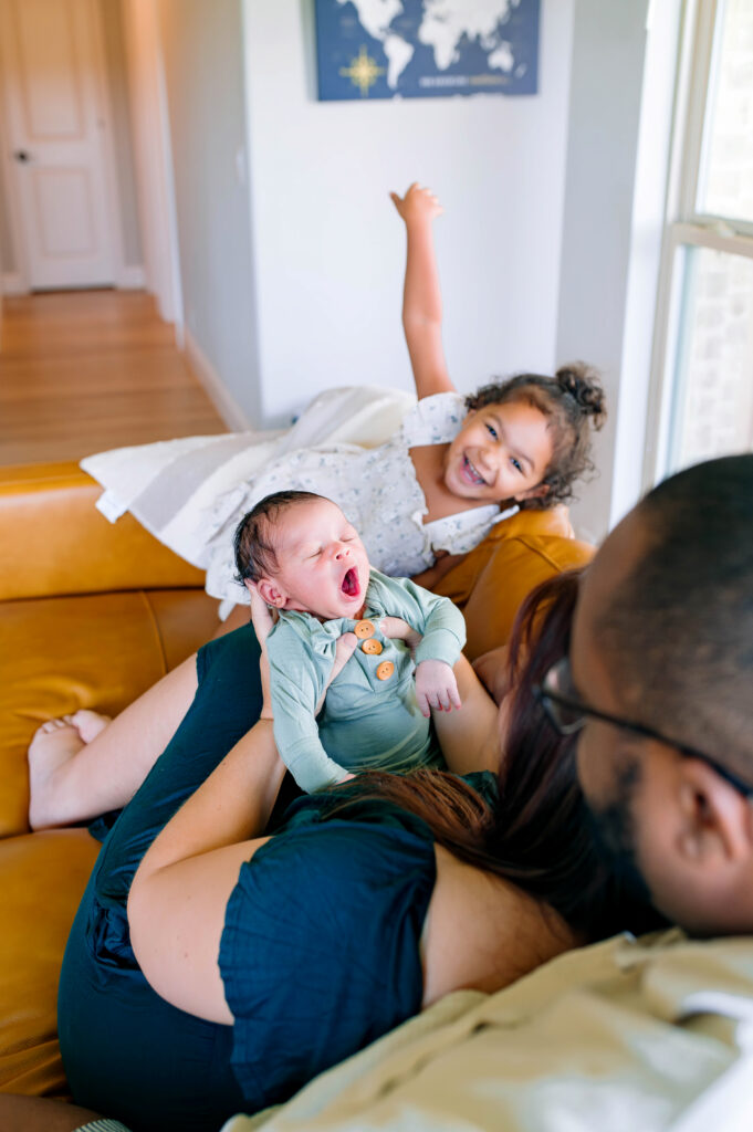 Lifestyle newborn photo of a baby resting on parents’ laps while an older sibling smiles nearby during a McKinney newborn photography session at home.