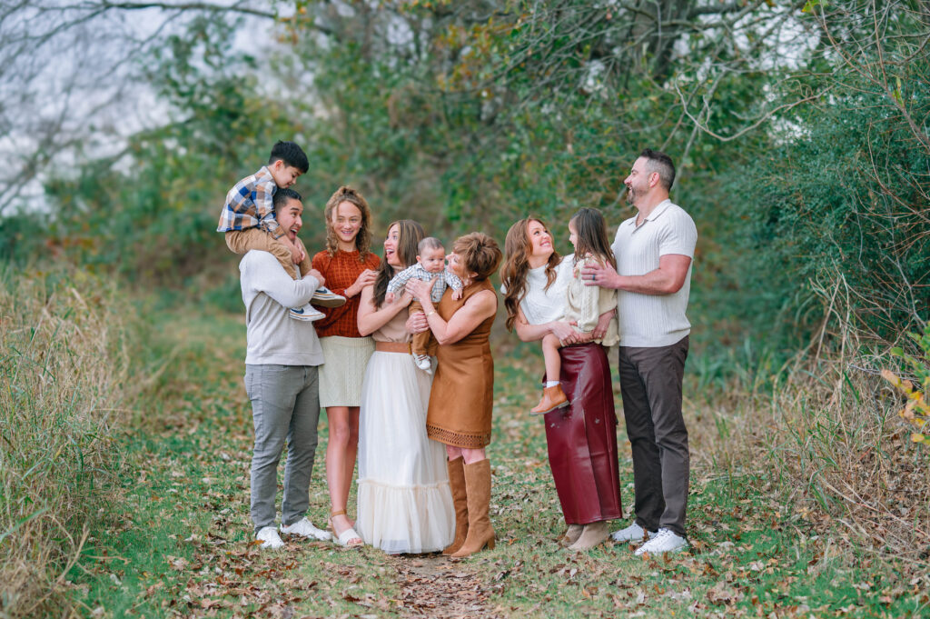 Extended family standing next to each other along a wooded trail at Erwin Park in McKinney, Texas, wearing coordinated fall outfits in warm neutral tones, smiling and laughing together during a holiday family session by a McKinney Extended Family Photographer.