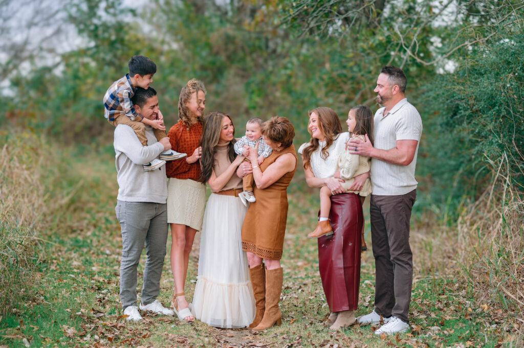 Extended family smiling at little baby boy in the middle at Erwin Park in McKinney, Texas, wearing coordinated fall outfits in warm neutral tones, smiling and laughing together during a holiday family session by a McKinney Extended Family Photographer.
