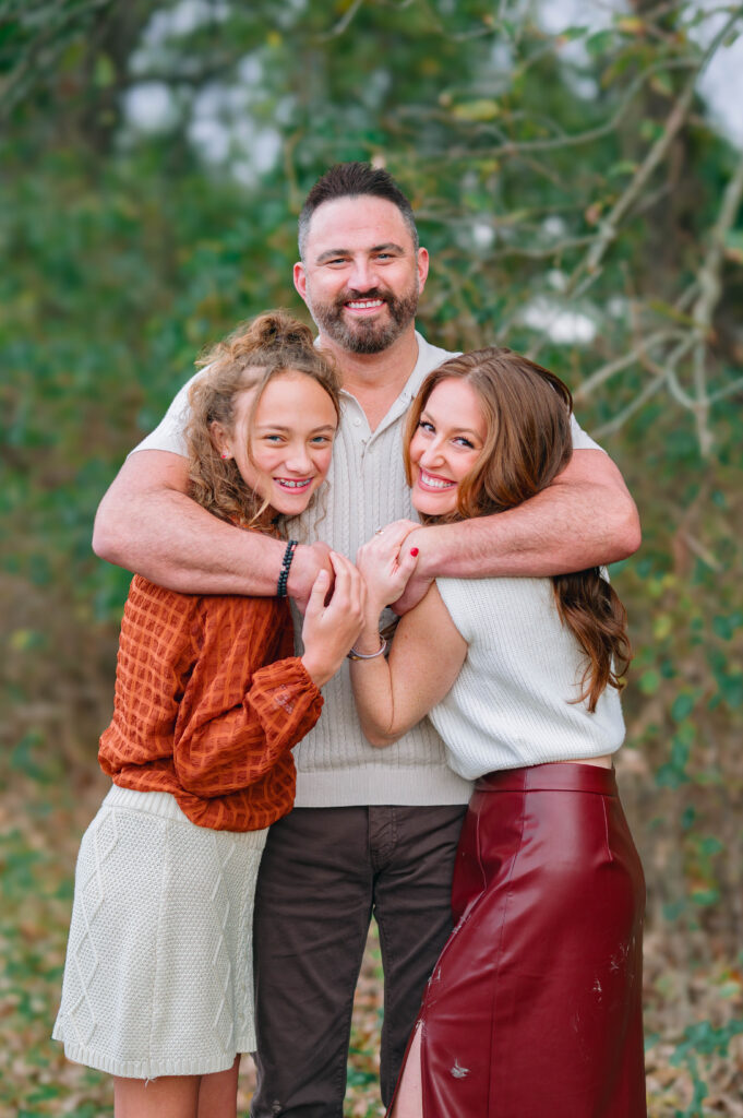 Dad hugging mom and daughter along a wooded trail at Erwin Park in McKinney, Texas, wearing coordinated fall outfits in warm neutral tones, smiling and laughing together during a holiday family session by a McKinney Extended Family Photographer.