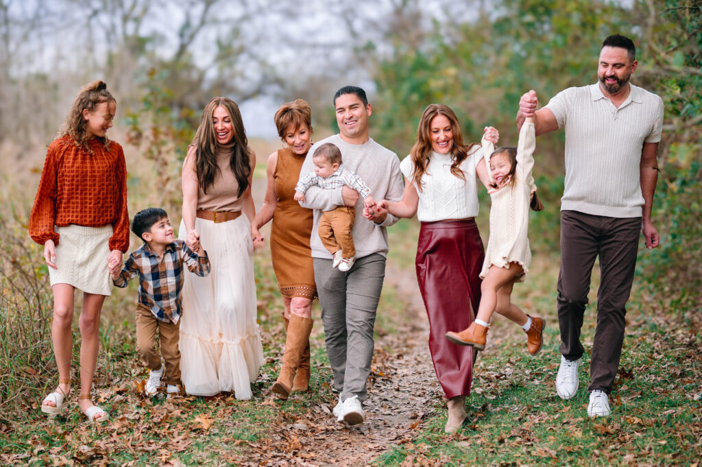 Extended family walking hand in hand along a wooded trail at Erwin Park in McKinney, Texas, wearing coordinated fall outfits in warm neutral tones, smiling and laughing together during a holiday family session by a McKinney Extended Family Photographer.