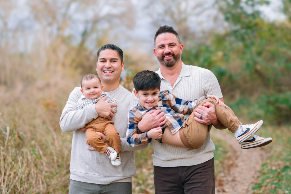 Two fathers holding two sons in arms while smiling along a wooded trail at Erwin Park in McKinney, Texas, wearing coordinated fall outfits in warm neutral tones, smiling and laughing together during a holiday family session by a McKinney Extended Family Photographer.