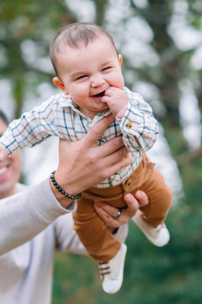 Dad holding up smiling baby boy along a wooded trail at Erwin Park in McKinney, Texas, wearing coordinated fall outfits in warm neutral tones, smiling and laughing together during a holiday family session by a McKinney Extended Family Photographer.