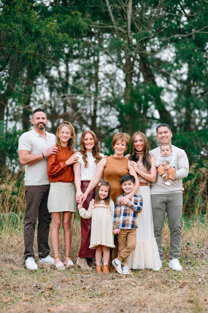 Extended family standing and smiling next to each other along a wooded trail at Erwin Park in McKinney, Texas, wearing coordinated fall outfits in warm neutral tones, smiling and laughing together during a holiday family session by a McKinney Extended Family Photographer.