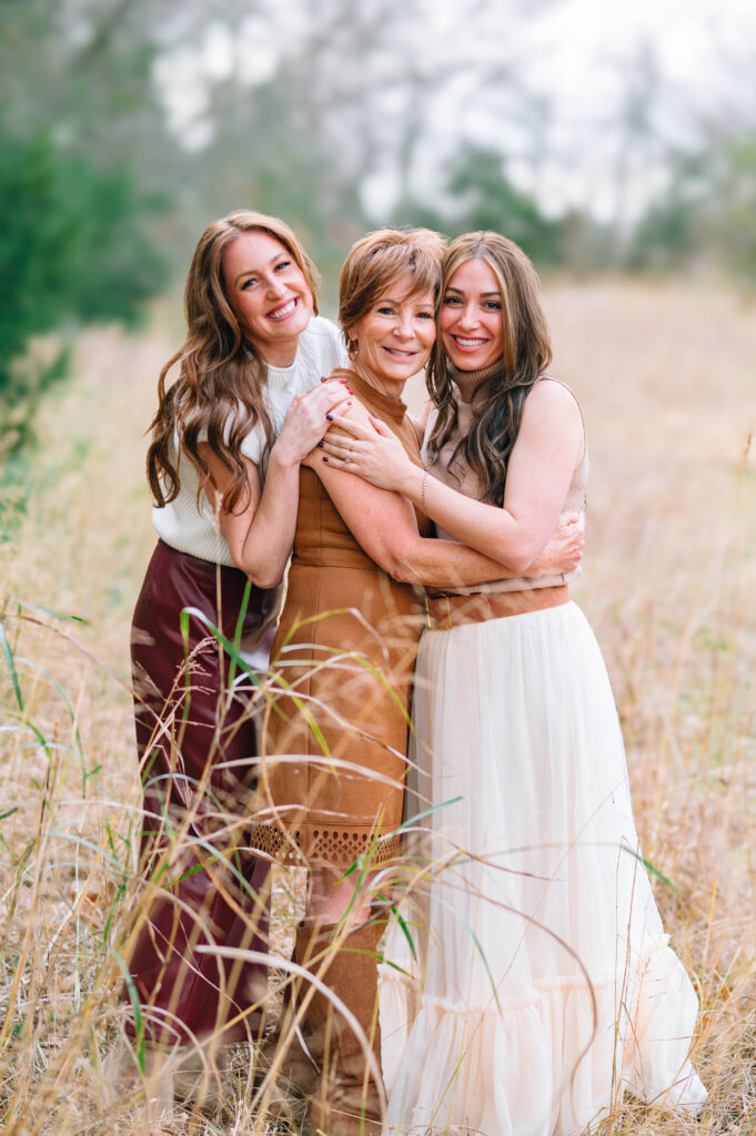 Three generations of women smiling and hugging in a grassy field at Erwin Park in McKinney, Texas, wearing coordinated neutral and fall-toned outfits during an extended family session by a McKinney Extended Family Photographer.