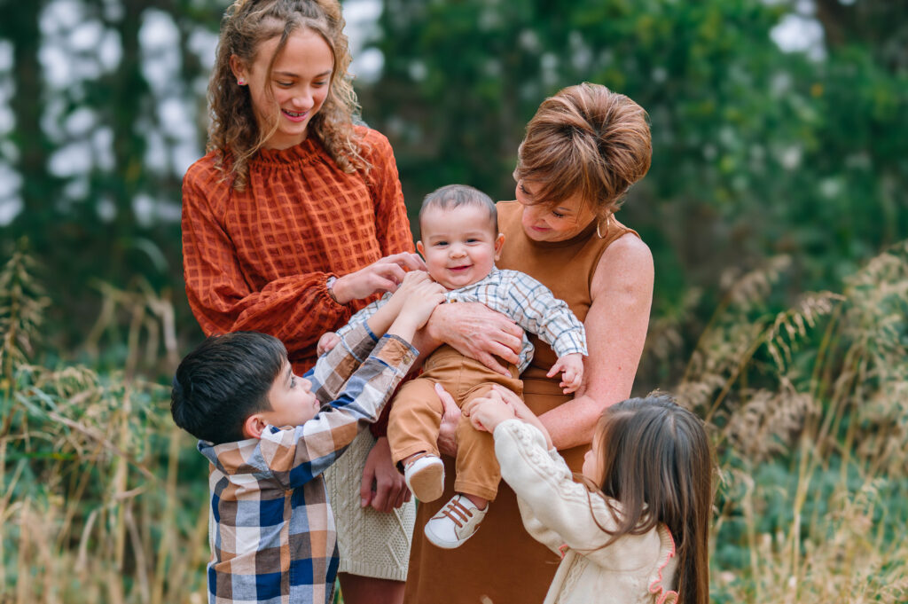 Grandmother standing with grandchildren holding baby grandson and all kids tickling baby in grandmas arm wooded trail at Erwin Park in McKinney, Texas, wearing coordinated fall outfits in warm neutral tones, smiling and laughing together during a holiday family session by a McKinney Extended Family Photographer.