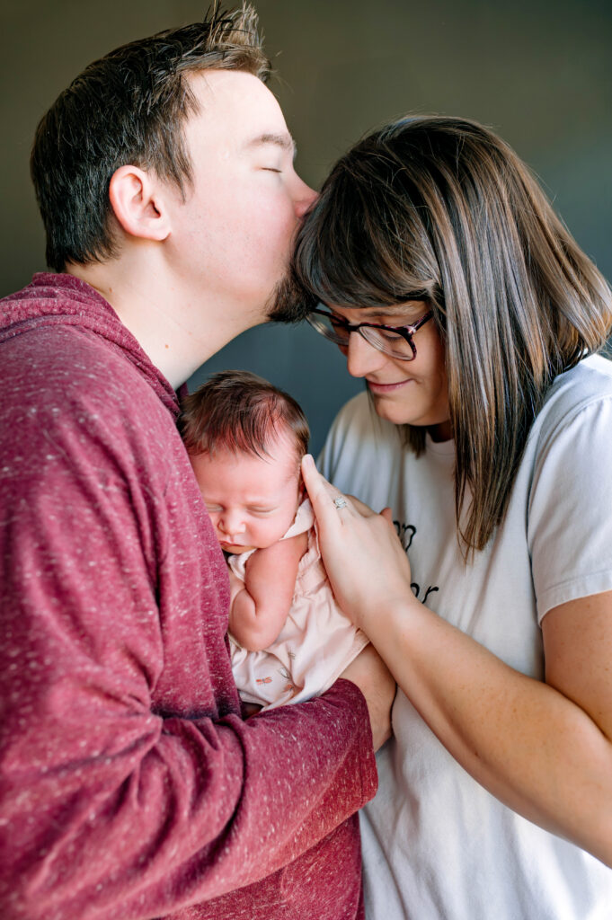 Mom and dad holding newborn daughter with mom looking at daughter with husband kissing moms head for their in home lifestyle newborn session in McKinney, Texas, by a McKinney Newborn Photographer.