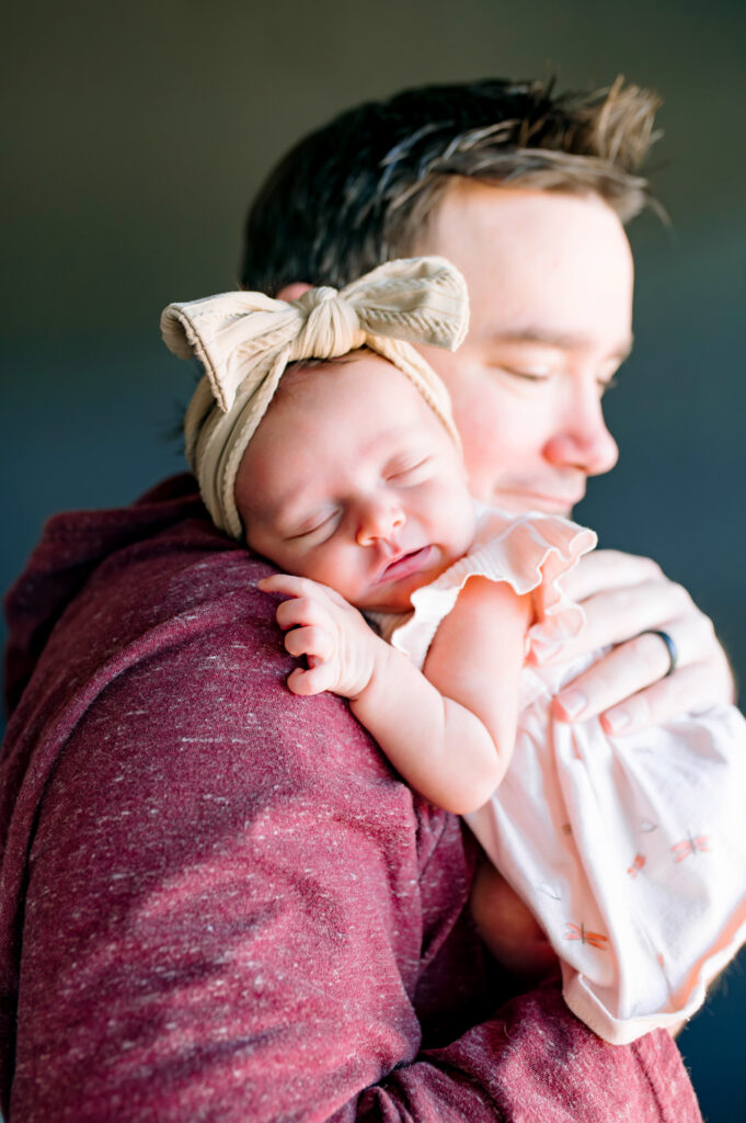 Dad holding newborn daughter asleep on his shoulder for his in home lifestyle newborn session in McKinney, Texas, wearing a maroon hoodie, by a McKinney Newborn Photographer.