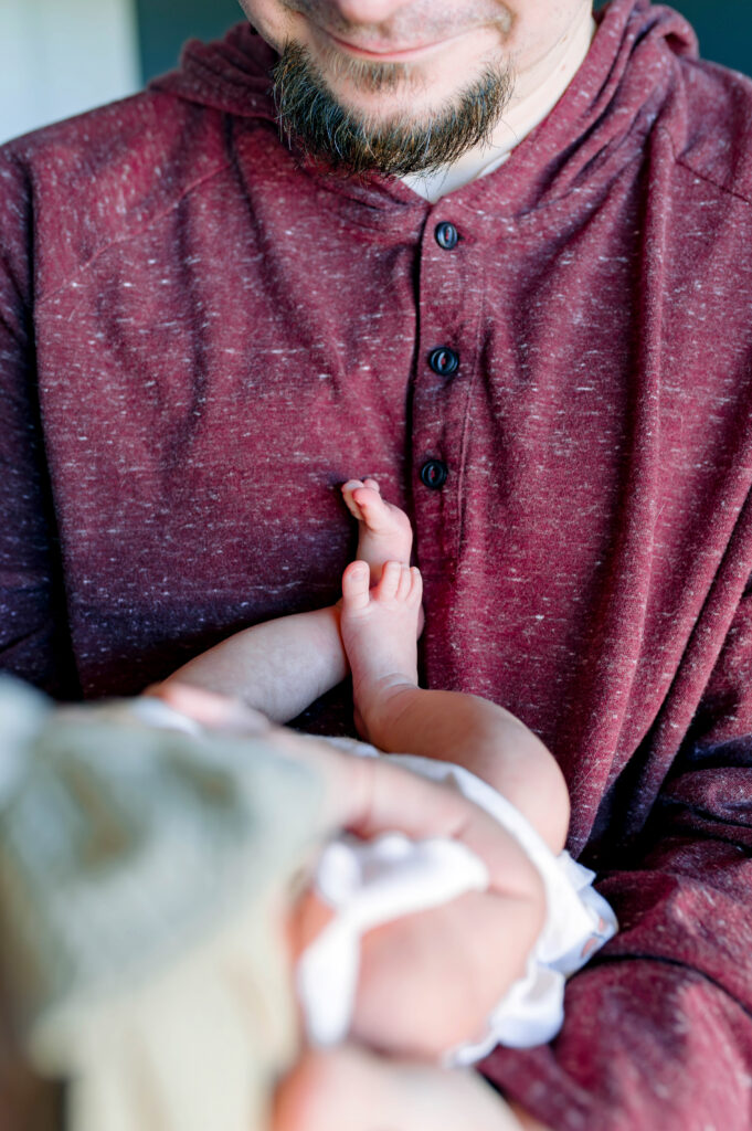 Dad holding newborn daughter looking at him with her feet showing for his in home lifestyle newborn session in McKinney, Texas, wearing a maroon hoodie, by a McKinney Newborn Photographer.