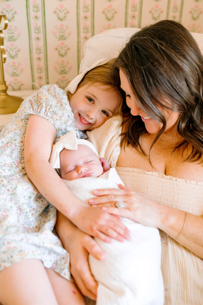 Baby girl in moms arms asleep with big sister laying on moms shoulder smiling at the camera during newborn session in Murphy, Texas. 