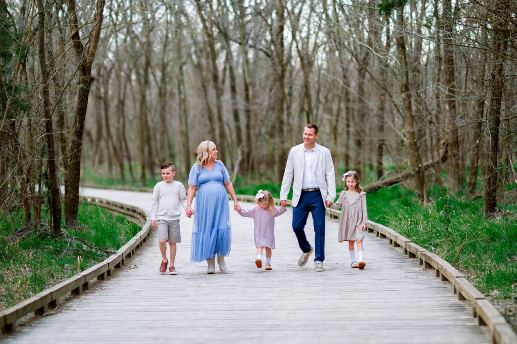 Family of five walking hand in hand on a wooden walkway at Oak Point Park in Plano, Texas, with three children between two parents and a pregnant mom glowing during a maternity session with Plano Maternity Photographer. 