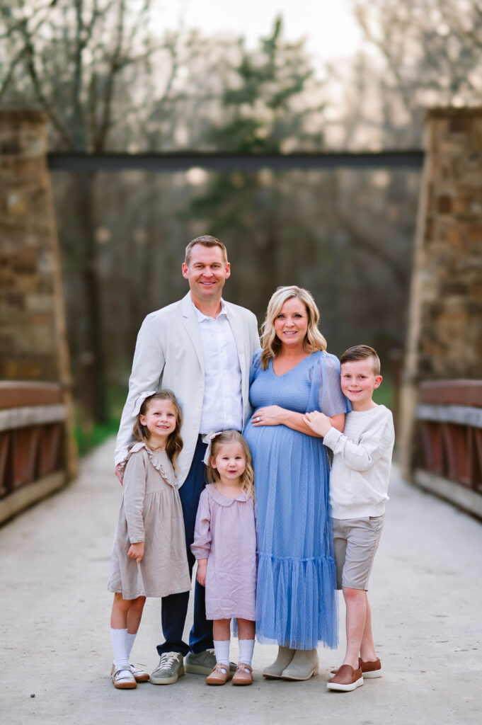 Family of 5 smiling and posing on bridge with pregnant mom at Oak Point Park in Plano, Texas, during a maternity session with Plano Maternity Photographer. 