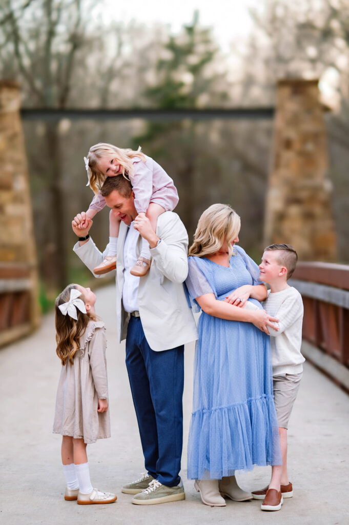 Family of 5 smiling at each other on bridge with pregnant mom at Oak Point Park in Plano, Texas, during a maternity session with Plano Maternity Photographer. 
