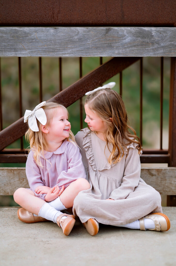 Sisters sitting and looking at each other on bridge at Oak Point Park in Plano, Texas, during a maternity session with Plano Maternity Photographer. 