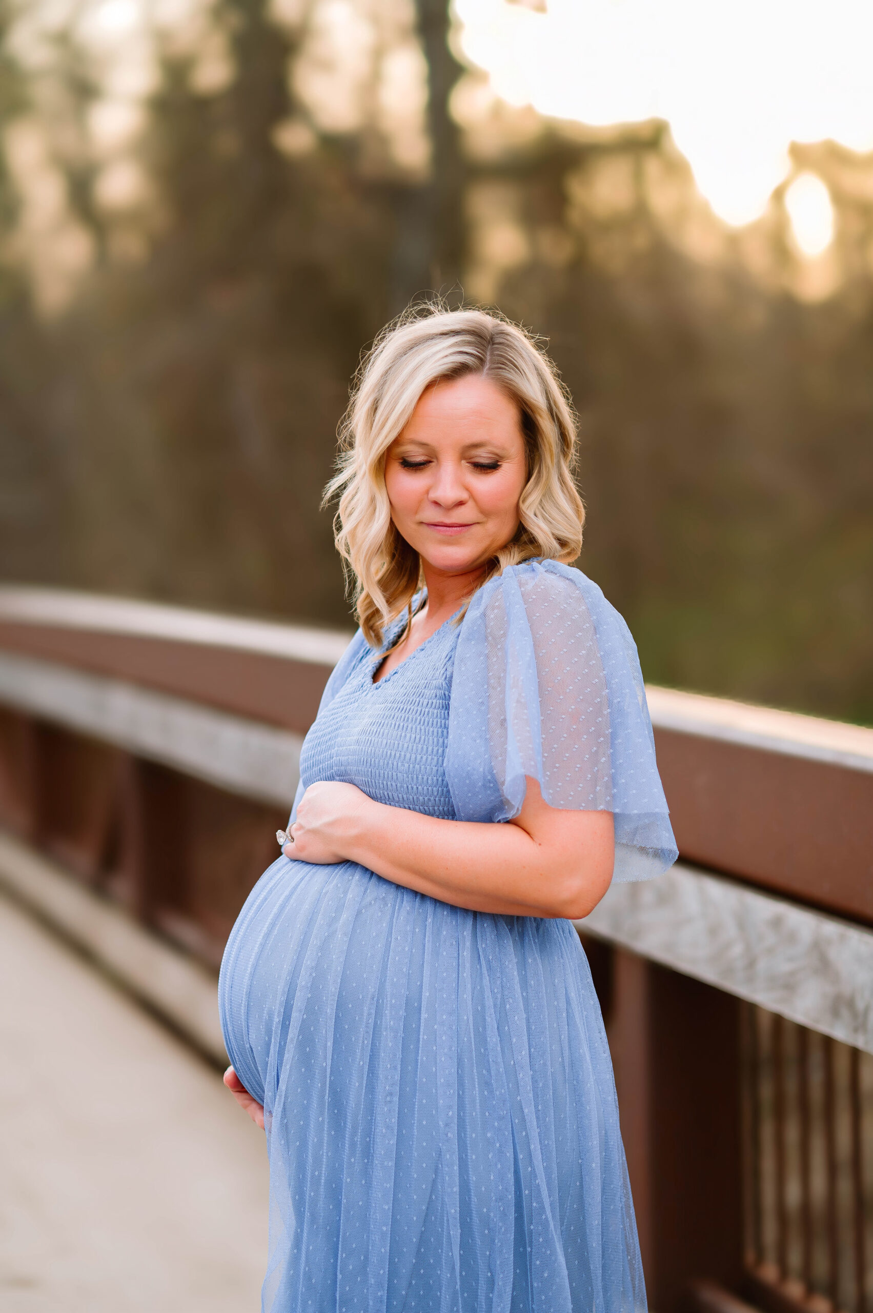 Pregnant mom looking down hugging her pregnant belly on bridge at Oak Point Park in Plano, Texas, during a maternity session with Plano Maternity Photographer.