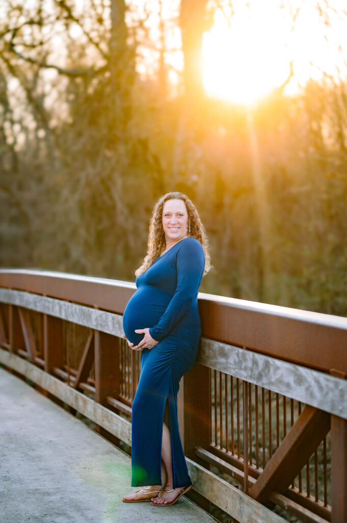 Plano maternity photographer capturing pregnant mom on bridge with sun glowing in the back at Oak Point Nature Preserve in Plano, Texas for a maternity session in Plano, Texas.