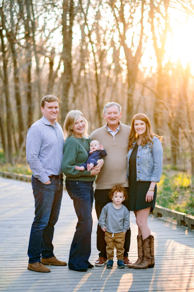 Grandparents holding baby grandson with toddler grandson standing next to them with mom and dad by their side on wooden path through woods at Oak Point Park in Plano, Texas with the sun glowing through the trees behind them during a family session with their Plano Family Photographer. 