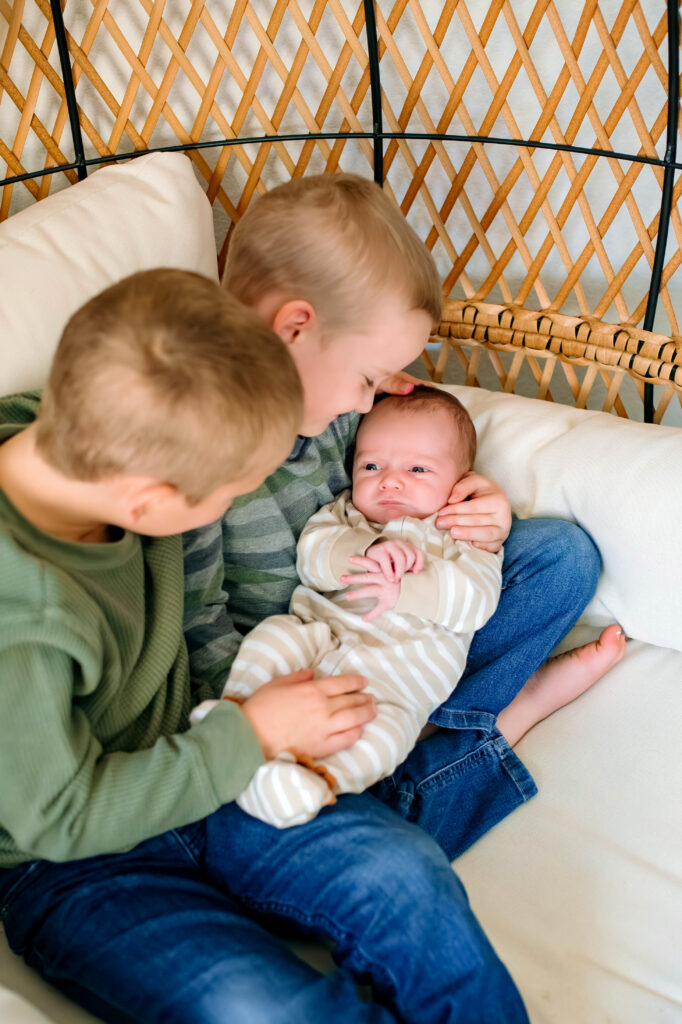 2 brothers hold baby brother in a wicker chair while baby is awake during a newborn session in Rockwall, Texas.