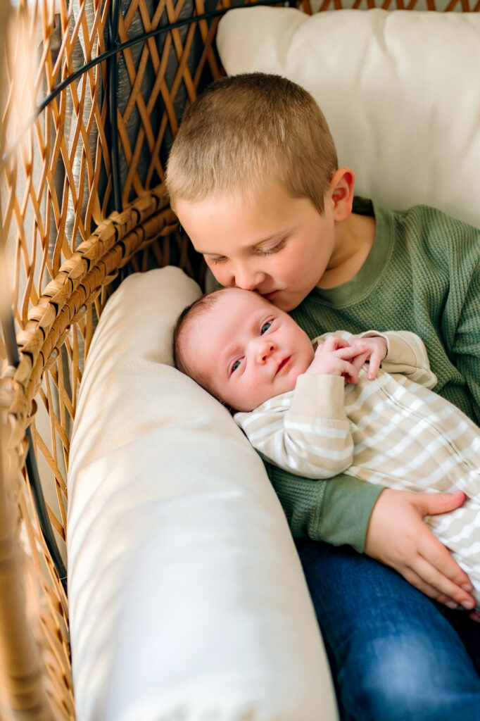 Big brother holding baby brother in arms while kissing babies head during newborn session in Rockwall, Texas.