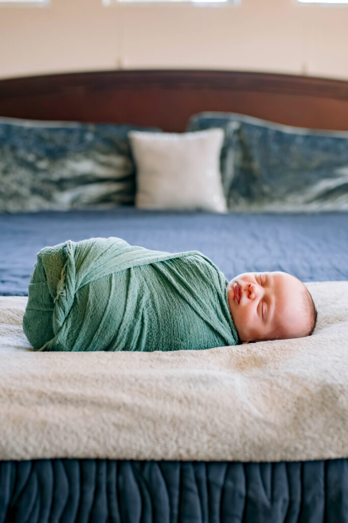 Baby boy wrapping in green on blanket on mom and dads bed asleep during newborn session in Rockwall, Texas.