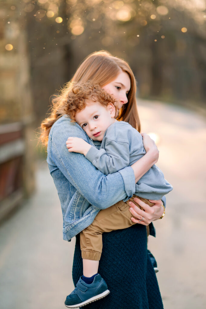 Son laying on mom's shoulder on bridge at Oak Point Park in Plano, Texas with the sun glowing behind them during a family session with their Plano Family Photographer. 
