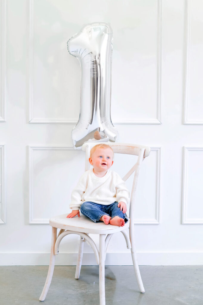 McKinney milestone photographer capturing a one year old boy sitting on white wood chair with a one balloon behind him for a studio milestone portrait.