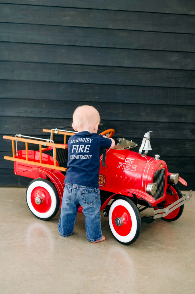 McKinney milestone photographer capturing a one year old boy standing in front of a small firetruck with a black wood wall behind him for a studio milestone portrait.