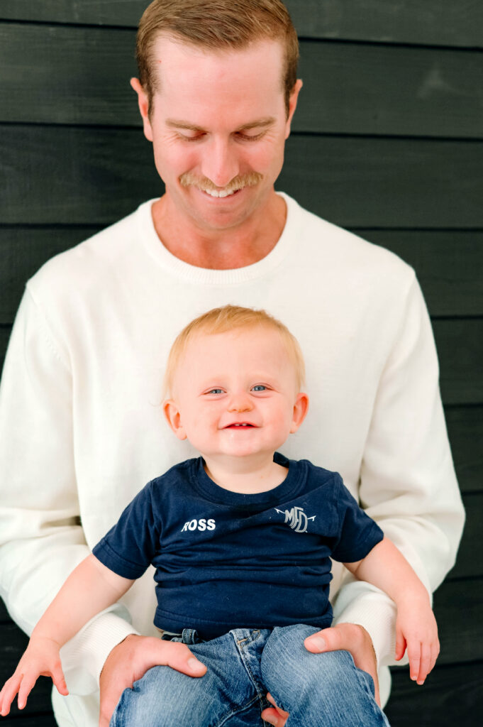 McKinney milestone photographer capturing a one year old boy in daddies arms facing out with a black wood wall behind him for a studio milestone portrait.