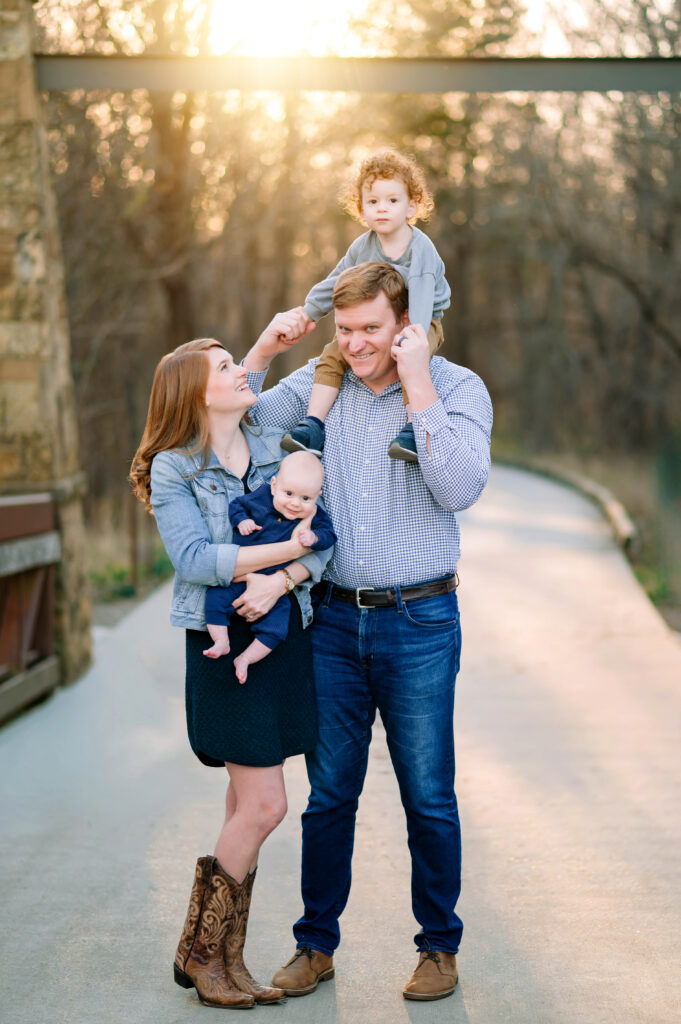 Mom looking up at toddler son on dad's shoulders while holding her smiling baby boy on bridge at Oak Point Park in Plano, Texas with the sun glowing behind them during a family session with their Plano Family Photographer. 