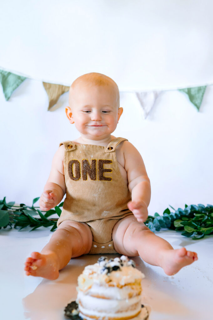 One year old little boy in overalls looking at his birthday cake with a white backdrop for his in home cake smash session in Frisco, Texas.