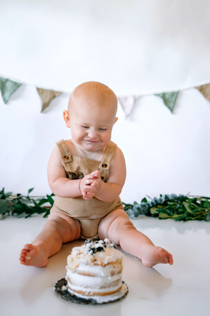 One year old little boy in overalls looking and smiling at his birthday cake with a white backdrop for his in home cake smash session in Frisco, Texas.