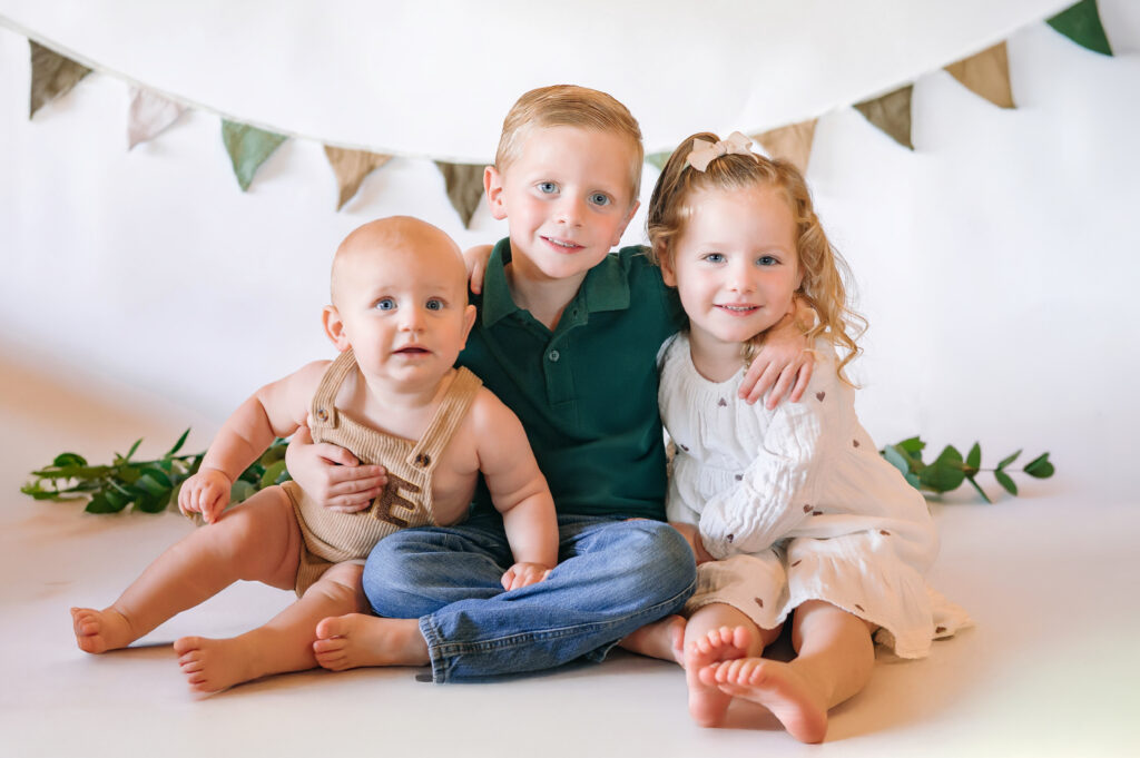 One year old little boy in overalls with older brother and sister with a white backdrop for his in home cake smash session in Frisco, Texas.