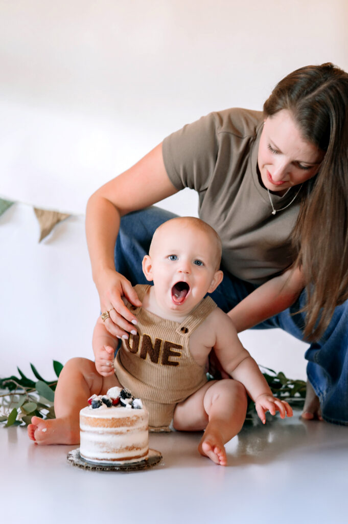 One year old little boy in overalls gagging over his birthday cake with mom behind him with a white backdrop for his in home cake smash session in Frisco, Texas.