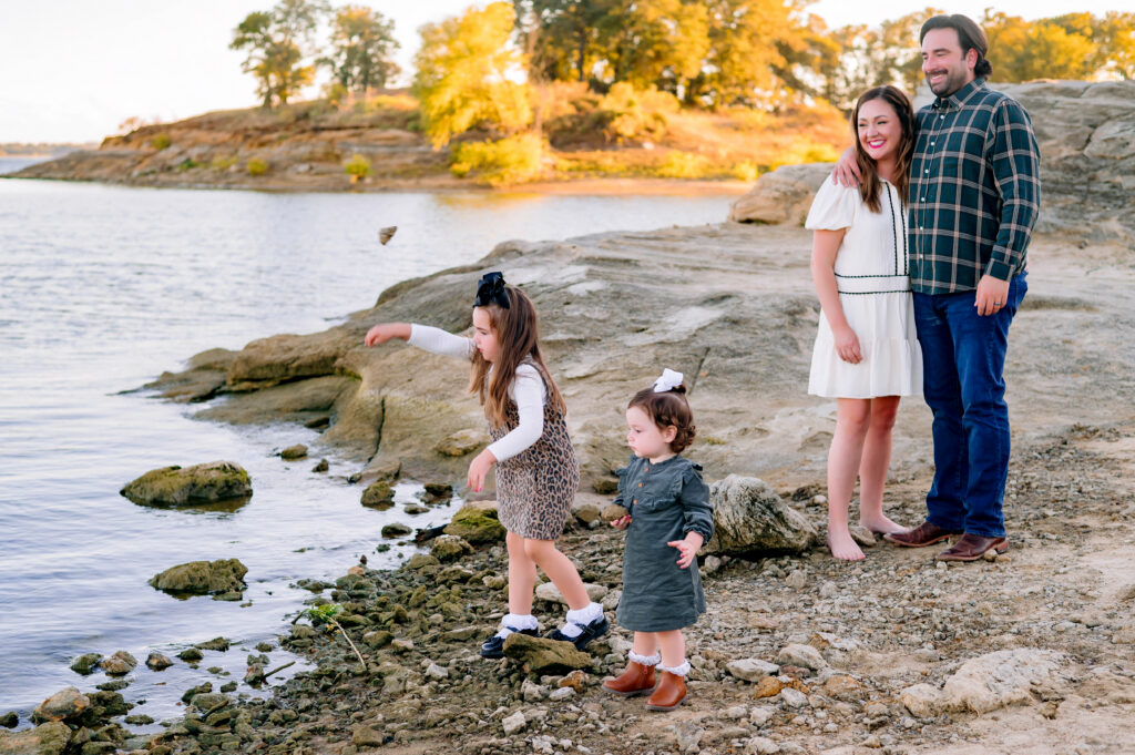 Mom and Dad watching little sisters throw rocks into the lake for a mini family session at Murall Park in Flower Mound Texas.