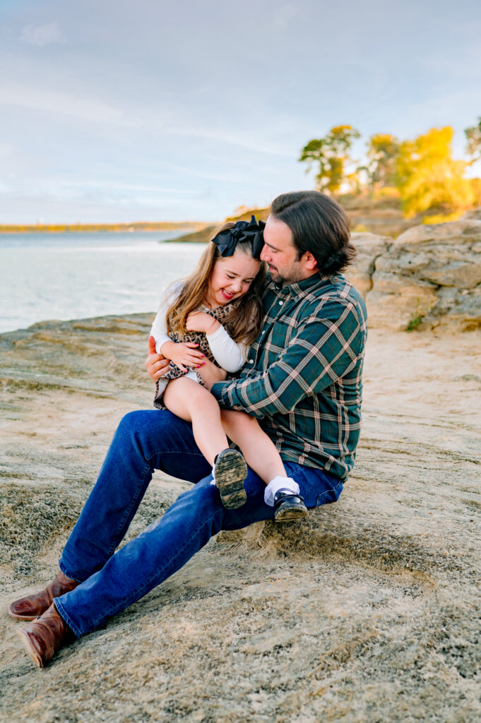 Daddy tickling daughter sitting on a rock for a mini family session at Murall Park in Flower Mound Texas.
