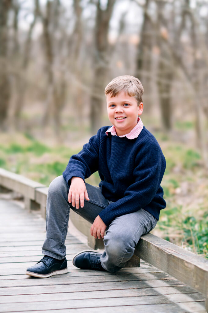 Little boy in jeans and navy sweater sitting on wood railing by wooden walkway at Oak Point Park in Plano, Texas with Jennifer L Kirk Photography.