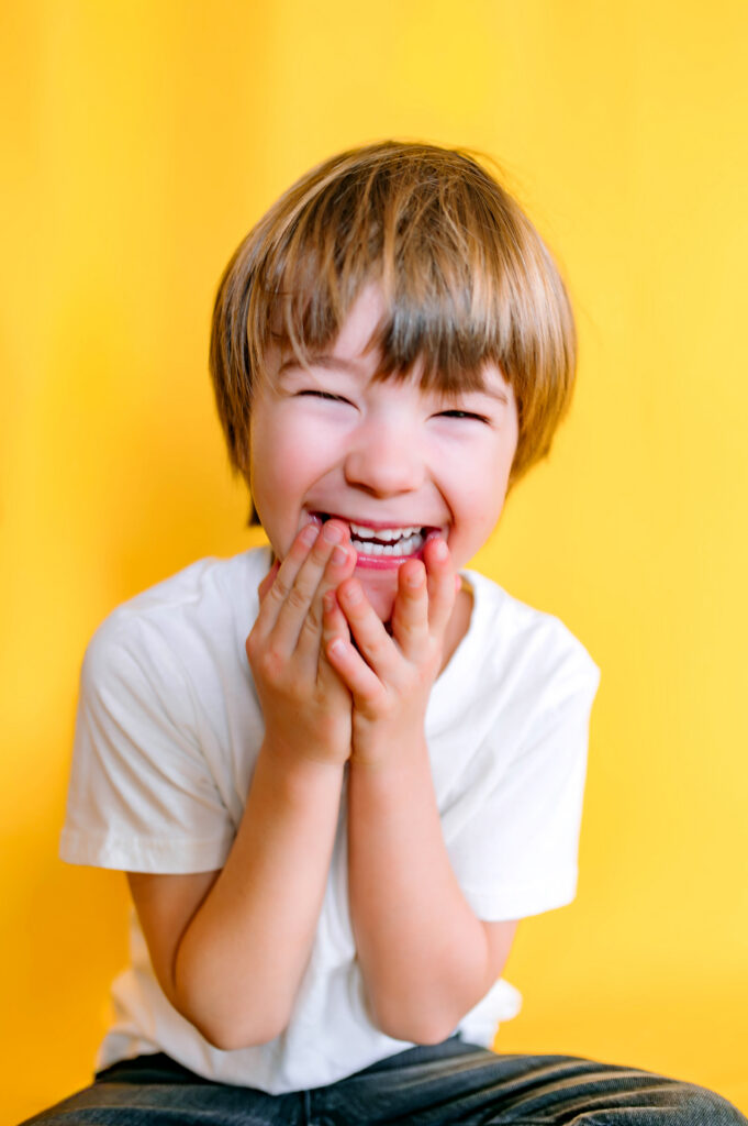 Little boy in white t-shirt laughing in front of yellow backdrop for personality pictures in Wylie, Texas. 