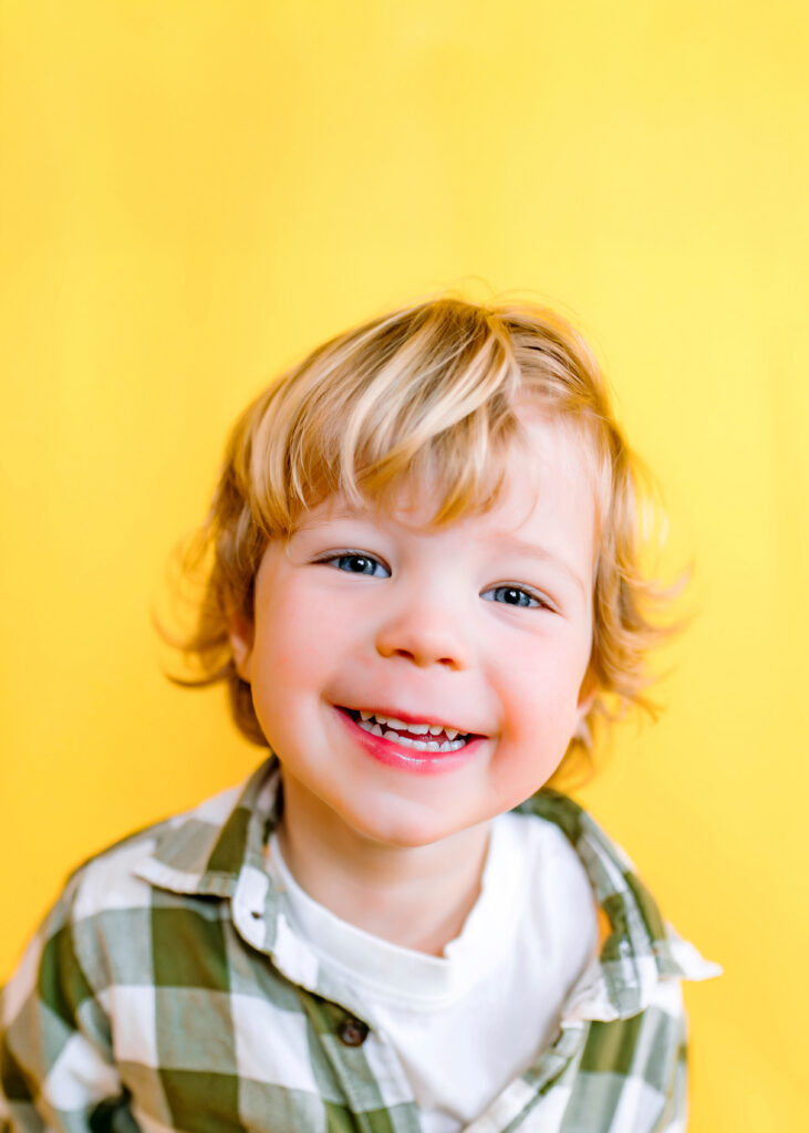 Little boy in green button up shirt smiling in front of yellow backdrop for personality pictures in Wylie, Texas with Jennifer L Kirk Photography. 