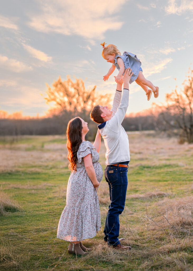 Dad throwing toddler daughter in the air with pregnant mom behind them looking at little girl in a field with sunset behind them during a maternity session in Plano, Texas with Jennifer L Kirk Photography. 