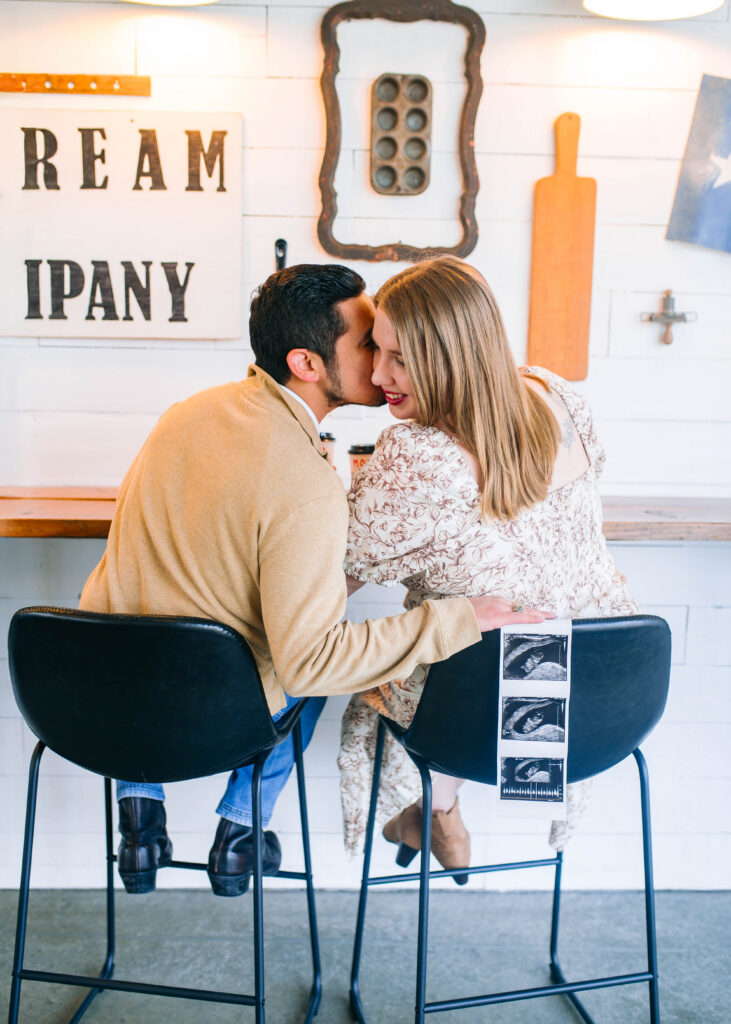 Couple sitting and man is whispering in girls ear at high bar chair next to a bar in a coffee shop with a sonogram hanging on the back of the chair during a birth announcement session in Rockwall, Texas. 