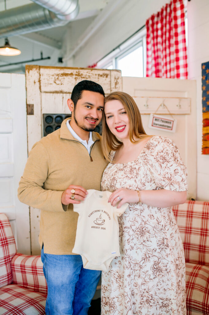 Mom and dad holding baby onesie in coffee shop during birth announcement photoshoot in Heath, Texas. 