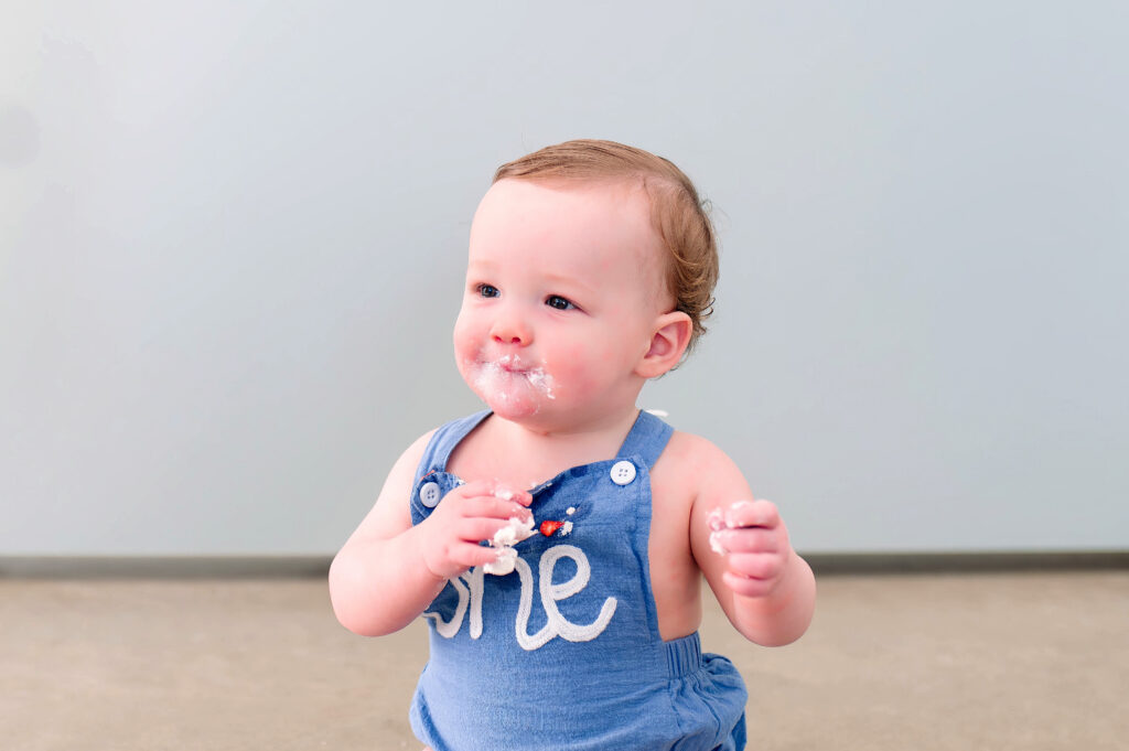 One year old boy sitting and smiling on concrete floor in front of a back drop during his milestone session at Lemondrop Studios in Mckinney, Texas. 