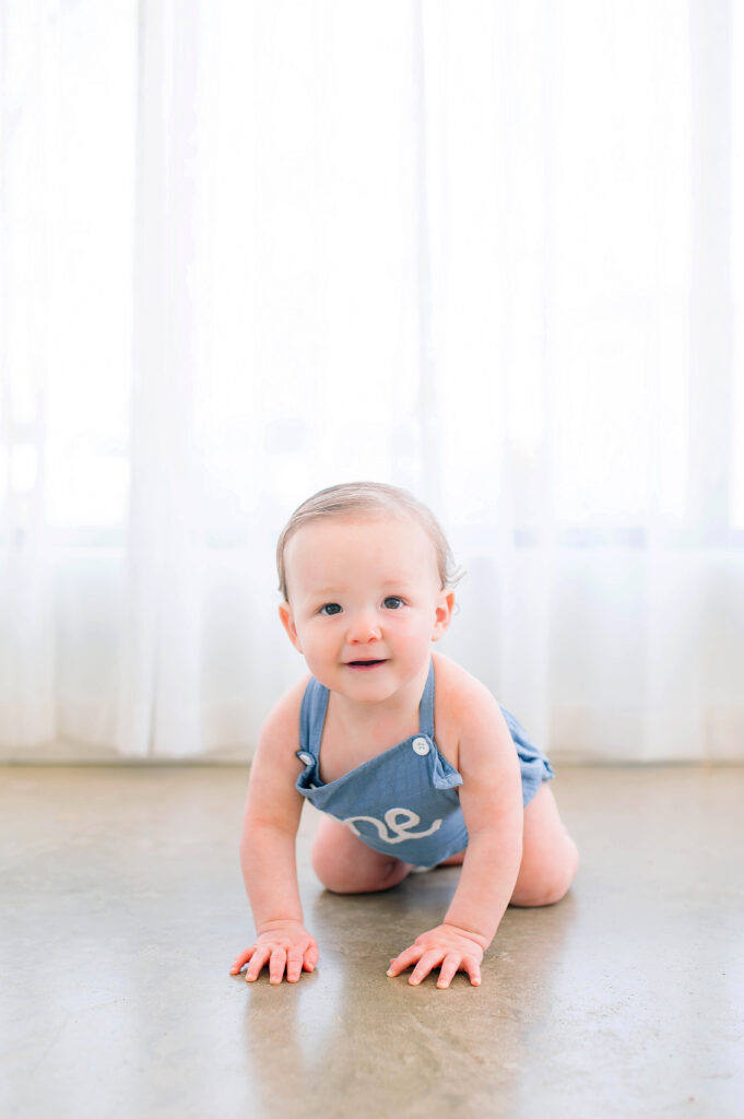 One year old boy crawling and smiling on concrete floor during his milestone session at Lemondrop Studios in Mckinney, Texas. 