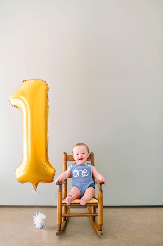 One year old boy sitting and smiling in wooden rocking chair next to a large gold 1 balloon during his milestone session at Lemondrop Studios in Mckinney, Texas. 