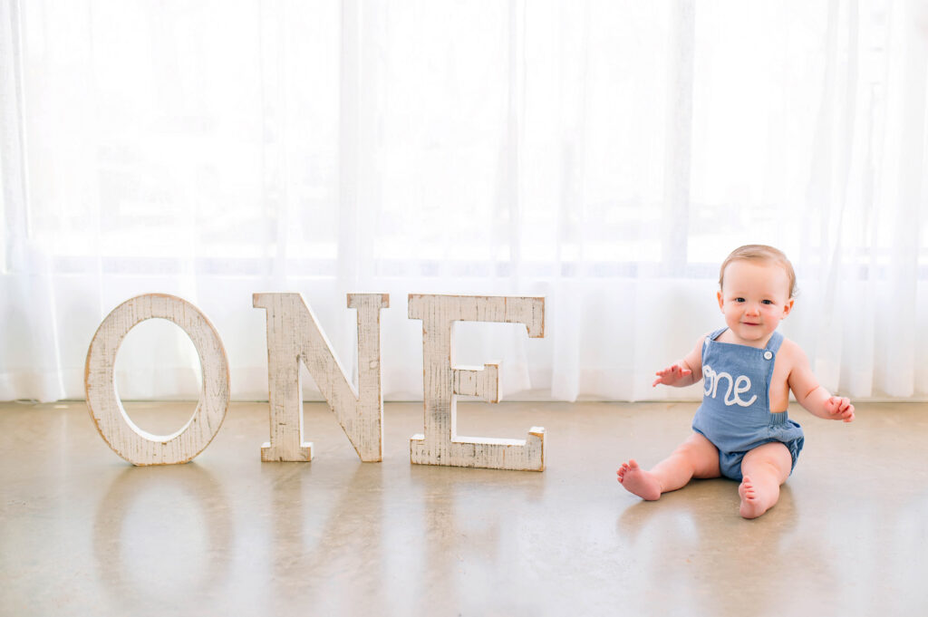 One year old boy sitting and smiling on concrete floor sitting next to the letters ONE during his milestone session at Lemondrop Studios in Mckinney, Texas. 