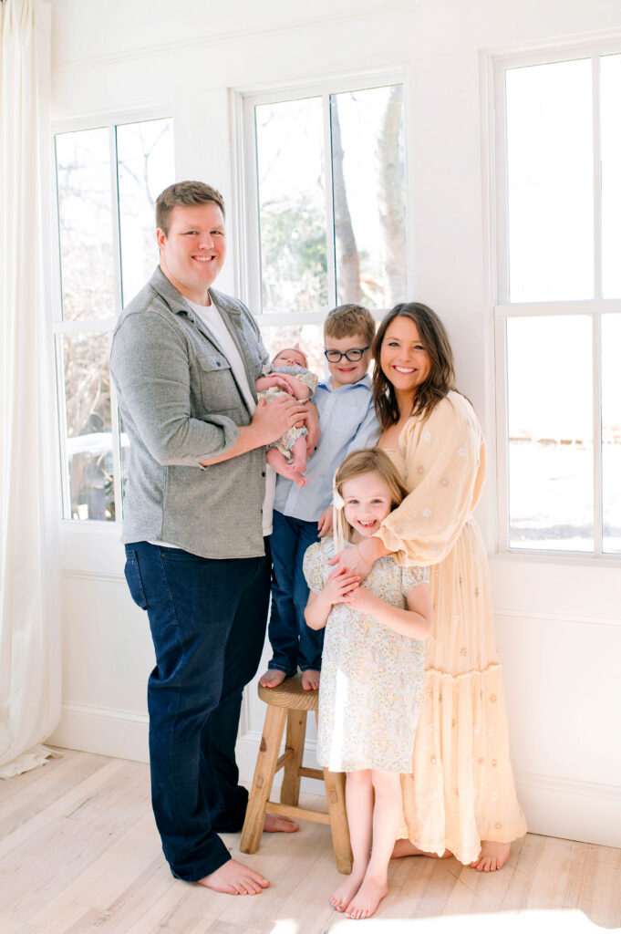 Family of 5 smiling and standing in front of a window during a in studio newborn session in Mckinney, Texas. 