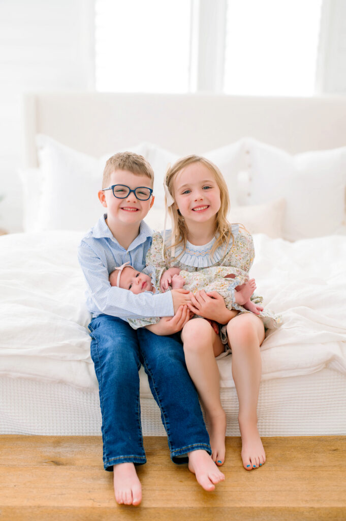 Big brother and big sister holding baby sister on a large bed smiling at the camera during their in studio newborn family session in Mckinney, Texas. 