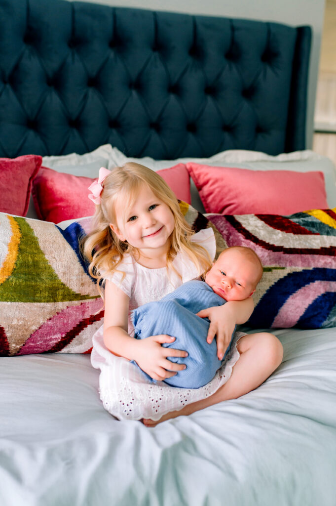 Little girl holding her baby brother on her parents bed smiling at the camera during a newborn session in Plano, Texas. 