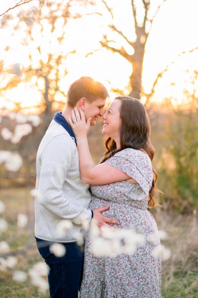 Pregnant mom and dad looking at each other behind a tree with flowers with the sunset behind them during a maternity session at Arbor Hill Nature Preserve in Plano, Texas. 